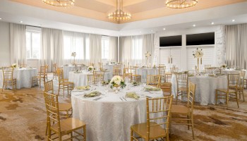 A decorated banquet hall with round tables, white tablecloths, floral centerpieces, and gold chairs, under elegant chandeliers.
