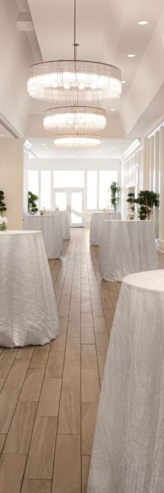 A hallway with round tables covered in white cloths, surrounded by potted plants and illuminated by chandeliers above.