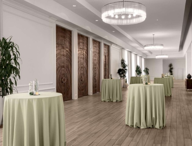 An elegant banquet hall with round tables covered in green cloth and large potted plants, featuring modern lighting and wooden doors.