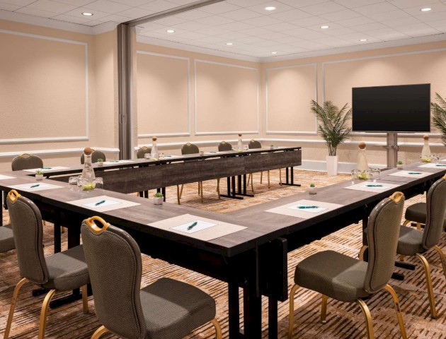 The image shows a conference room with a U-shaped table setup, chairs, notepads, pens, and a screen, decorated with plants.