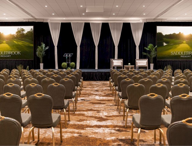 A conference room with rows of chairs facing a stage, featuring large screens displaying 