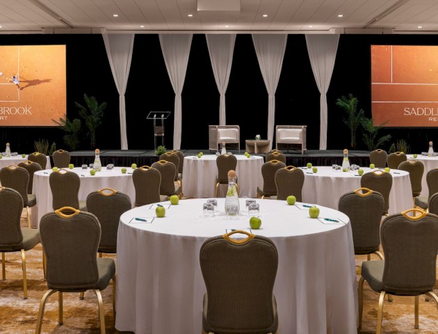 The image shows a conference room with round tables and chairs, a stage with a lectern, screens displaying 