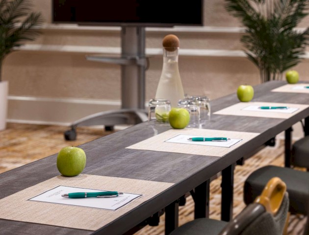 A conference table setup with notepads, pens, green apples, water glasses, a carafe, and plants in the background, ready for a meeting.
