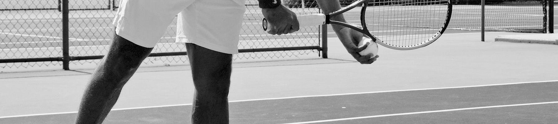 A person is on a tennis court preparing to serve, holding a racket, and wearing athletic clothing. The scene is outdoors in black and white.