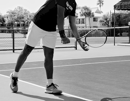 A person is on a tennis court preparing to serve, holding a racket, and wearing athletic clothing. The scene is outdoors in black and white.