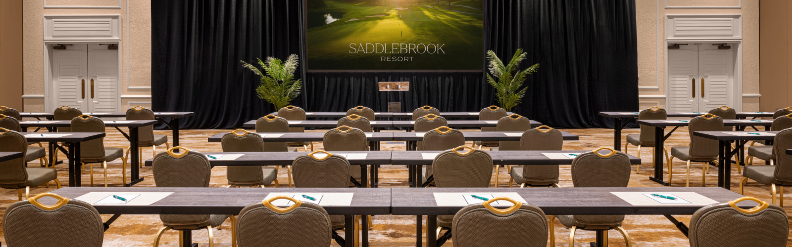 The image shows a conference room with rows of tables and chairs facing a large screen displaying 