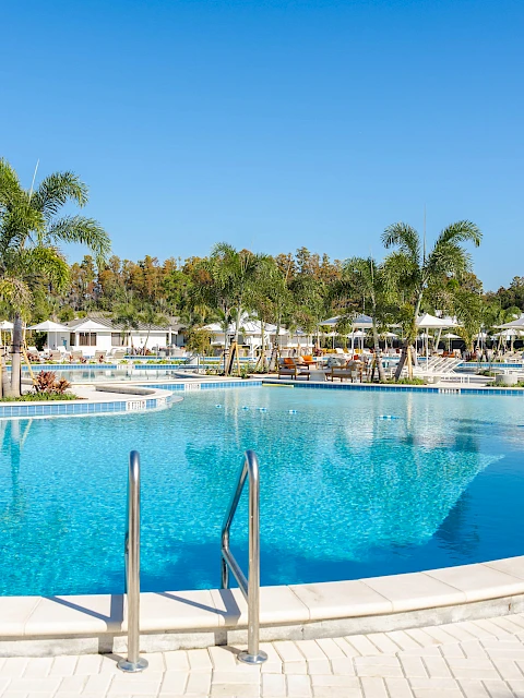 A luxurious outdoor pool area with palm trees, white lounge chairs, umbrellas, and cabanas under a bright blue sky.