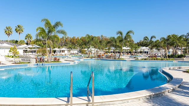 A luxurious outdoor pool area with palm trees, white lounge chairs, umbrellas, and cabanas under a bright blue sky.