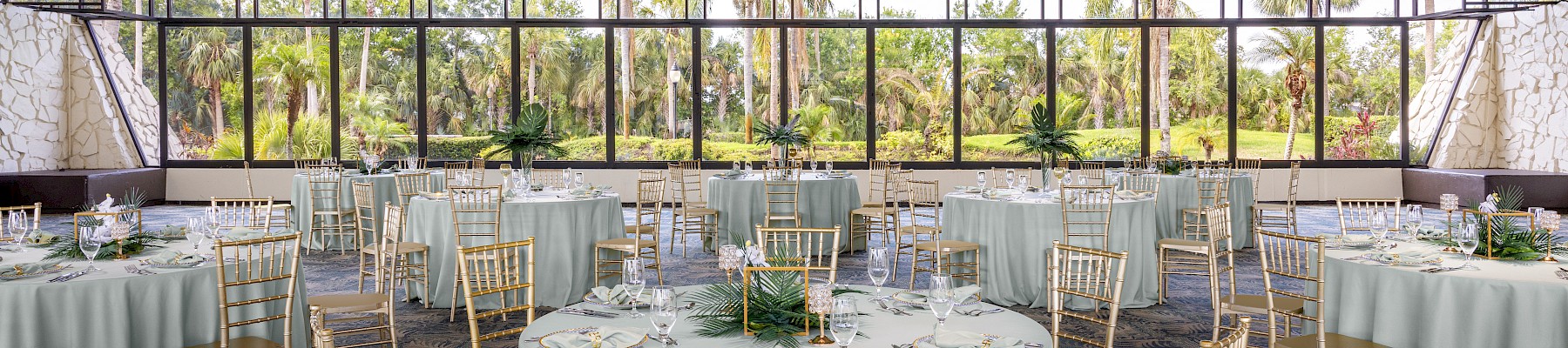 A decorated banquet hall with round tables, gold chairs, white tablecloths, and large windows revealing a lush, green outdoor view.