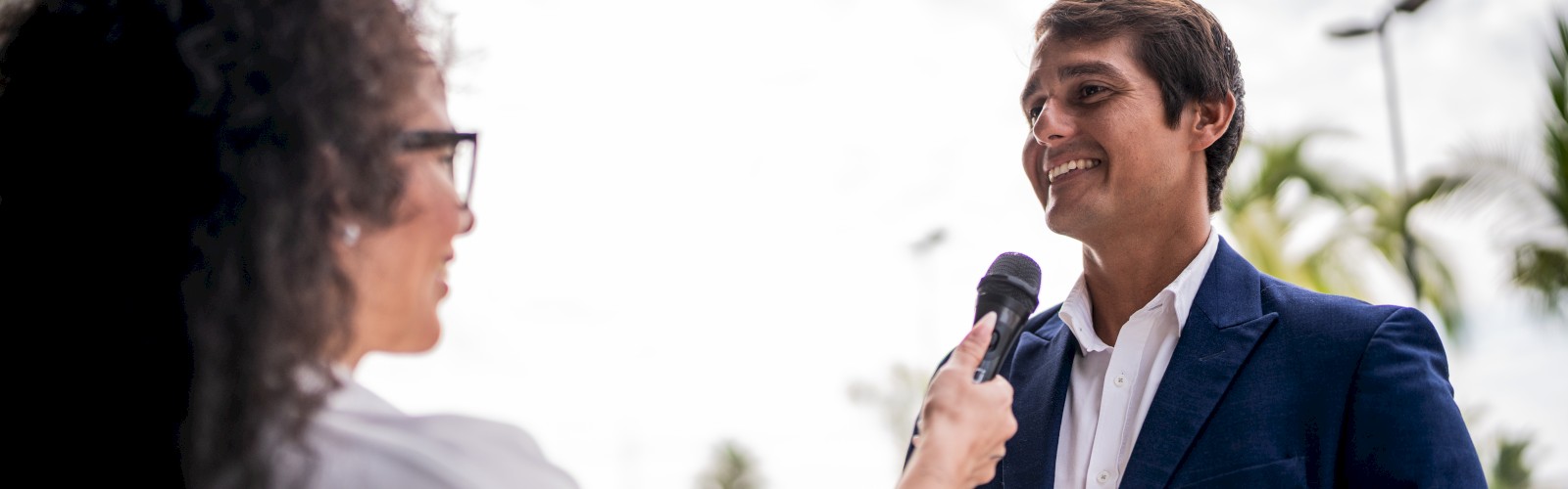 A woman is holding a microphone, interviewing a smiling man in a suit, outside on a cloudy day.