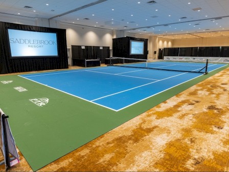 An indoor room transformed into a tennis court with blue and green flooring, screens displaying 