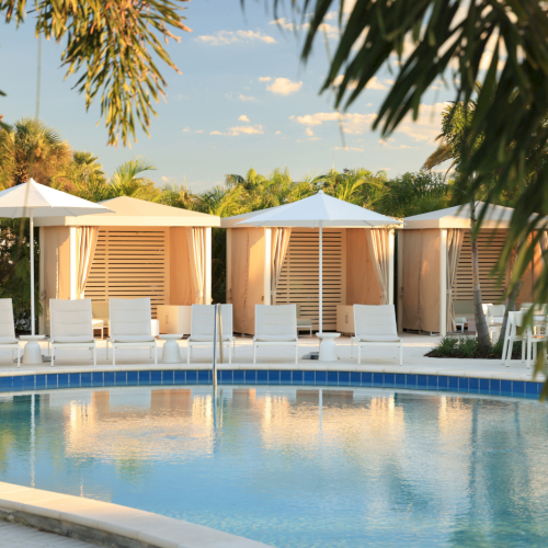 Luxurious poolside cabanas with white lounge chairs and umbrellas, surrounded by tropical greenery under a clear, sunny sky.