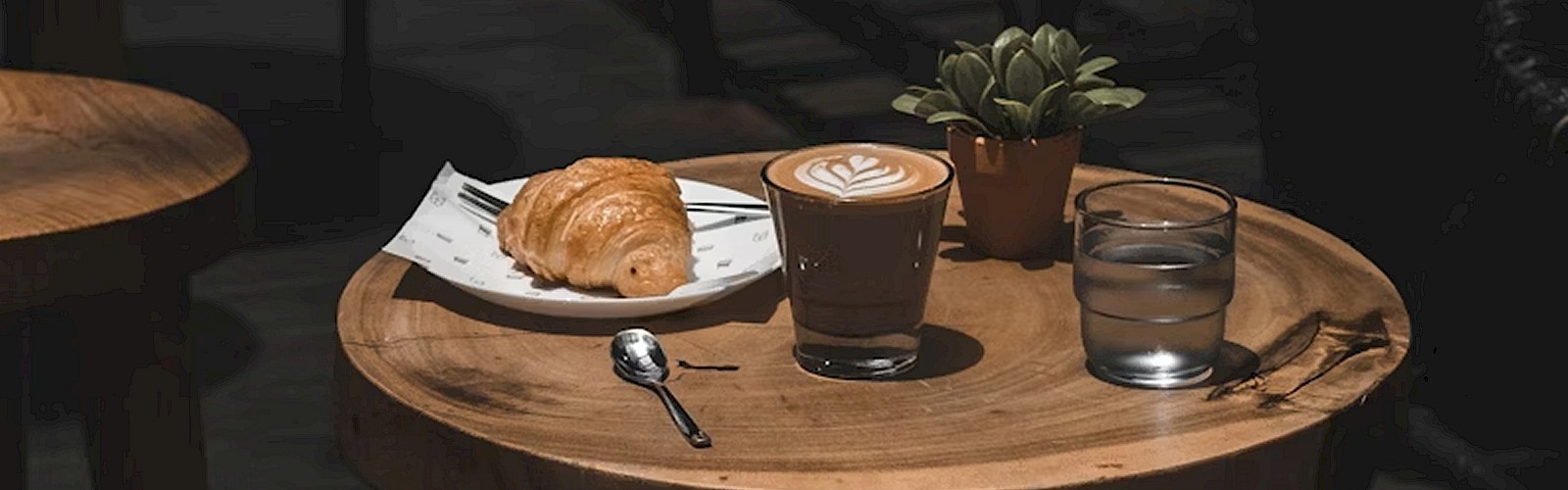 A wooden table with a croissant, coffee, water, and a small plant; an open book on a nearby chair, in a cozy café setting.