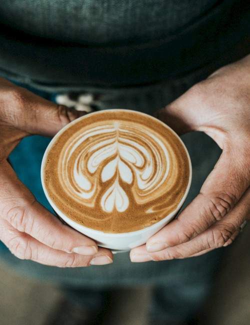 A person holds a cup of coffee with intricate latte art resembling a heart and leaf pattern in the foam on top.
