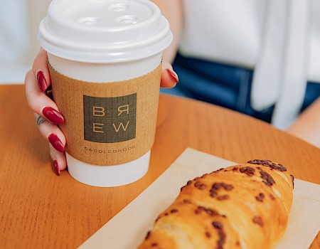A person with red nails holds a coffee cup while a pastry sits on a napkin beside it, both on an orange table.