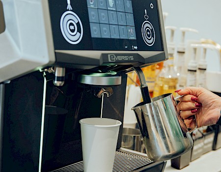 A modern espresso machine with digital interface, brewing a drink into a metal pitcher, while a barista prepares milk foam and a cup sits beneath.