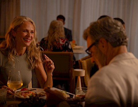 A woman with blonde hair smiles at a man across a dinner table, drinks and food in front of them, warm, intimate restaurant setting.
