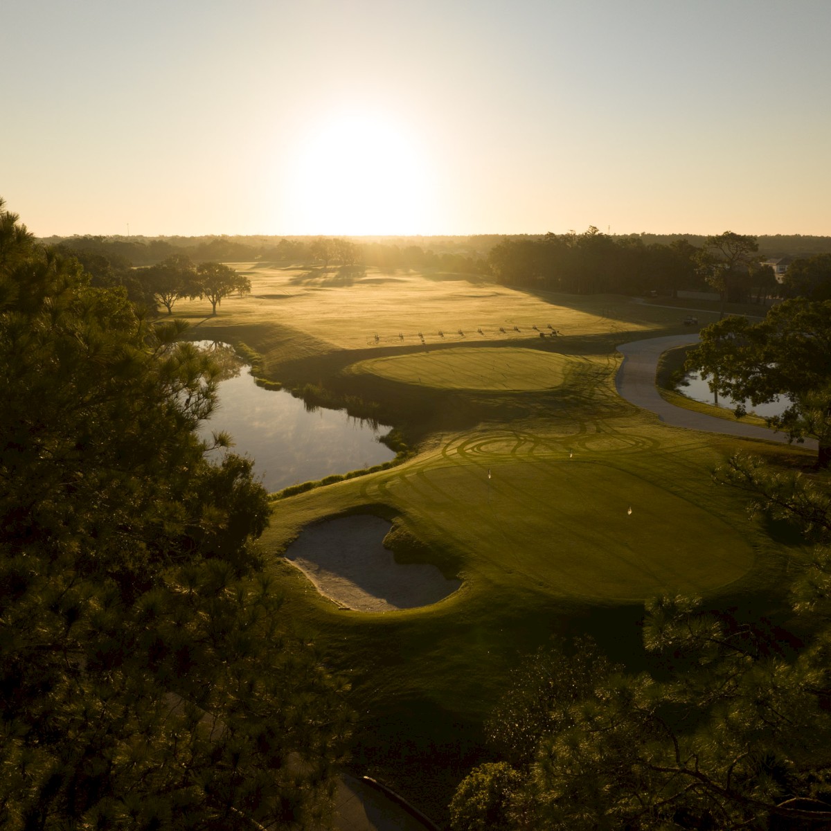 A scenic golf course at sunrise, featuring sand traps, water hazards, trees, and a winding path through lush greenery.