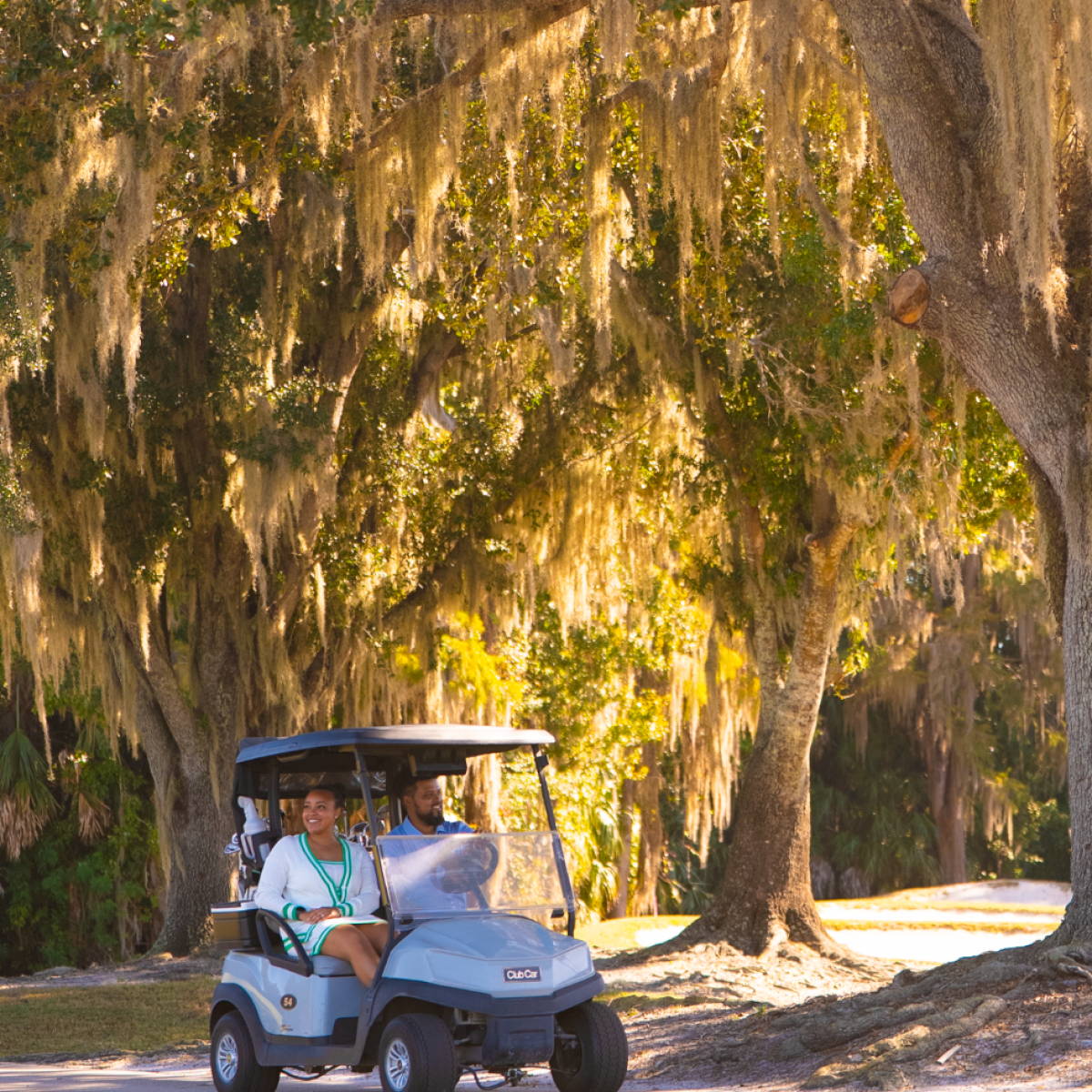 Two people are riding in a golf cart along a shaded path under large, moss-draped trees on a sunny day.