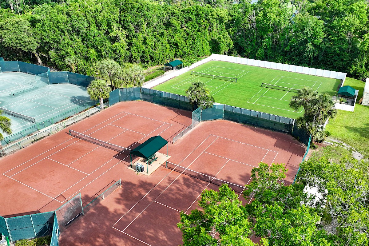 The image shows outdoor tennis courts with different surfaces, surrounded by greenery and trees, under a clear sky.