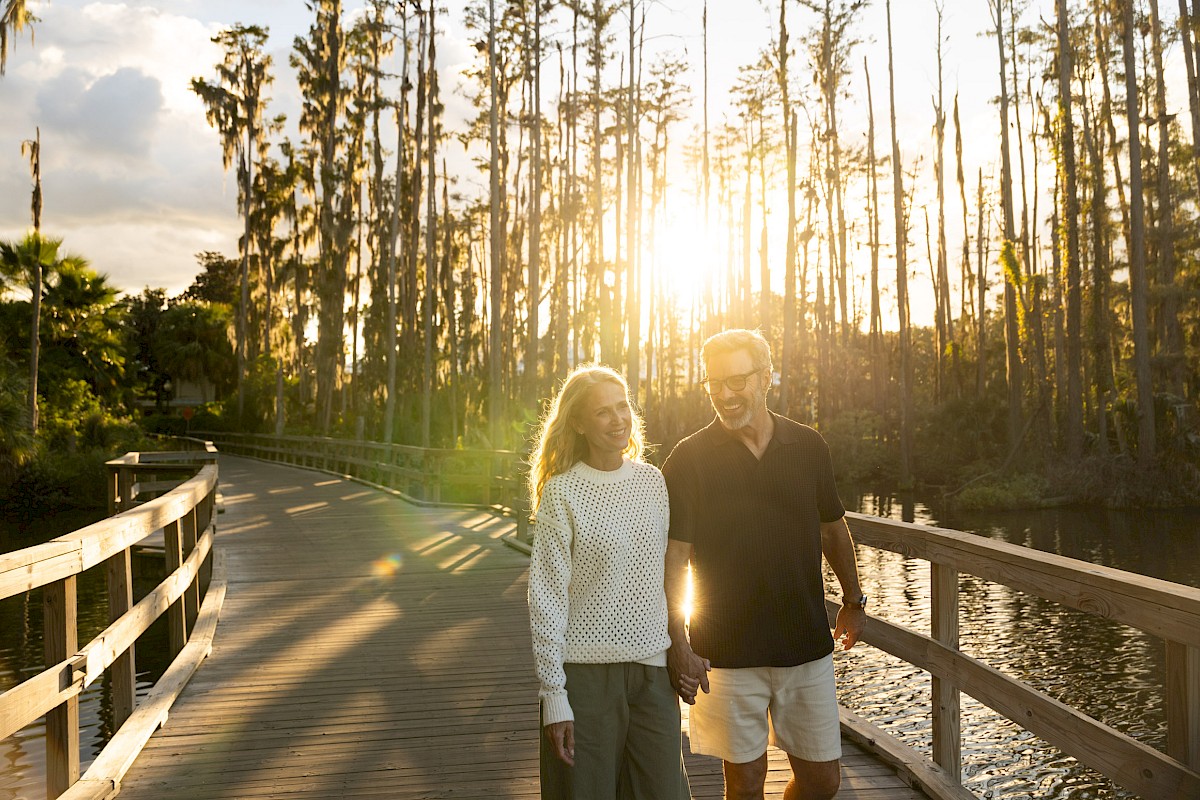 A couple walks on a sunlit wooden bridge through tall trees, warm sunset glow, peaceful nature scene, smiling and enjoying the moment.