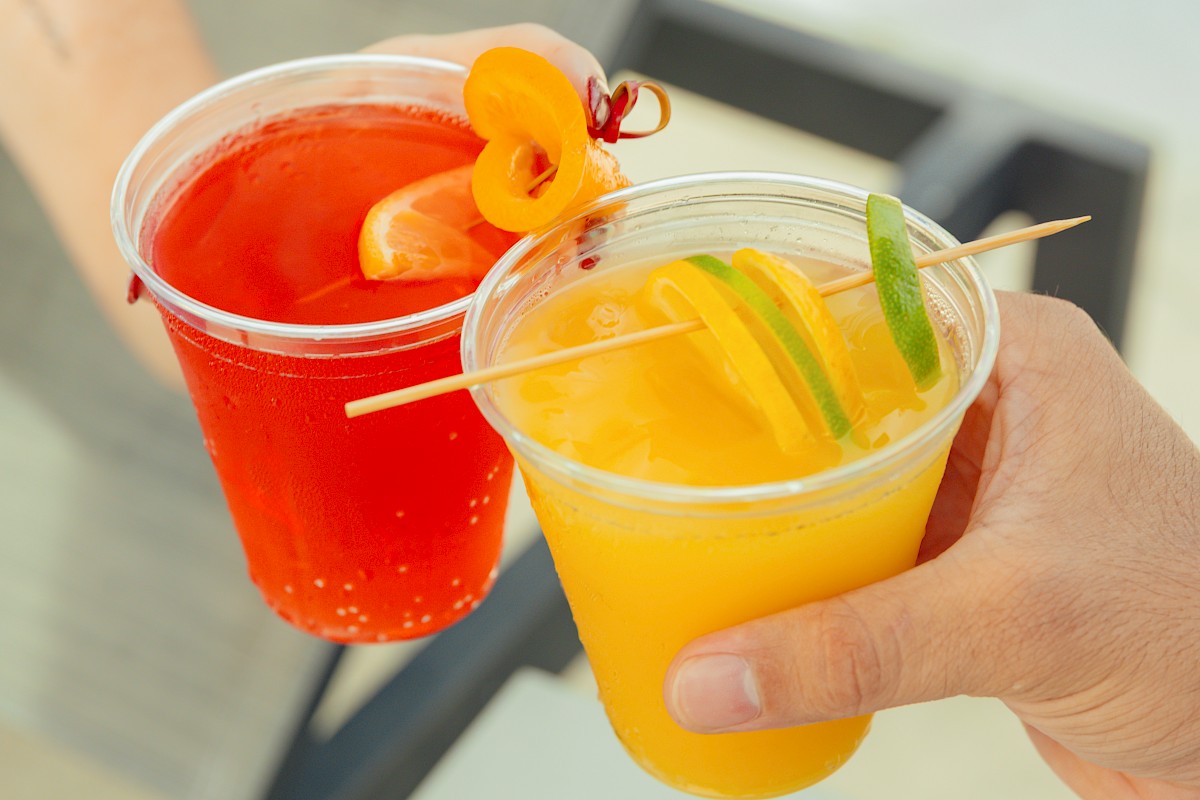 Two people are holding plastic cups with vibrant drinks, one red with an orange garnish and one yellow with lime slices.