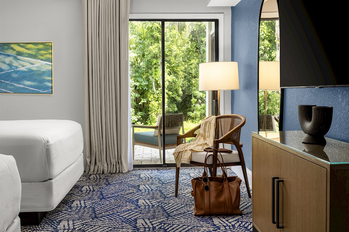 A hotel room with a bed, chair, lamp, and TV, featuring a blue accent wall and a window view of greenery.
