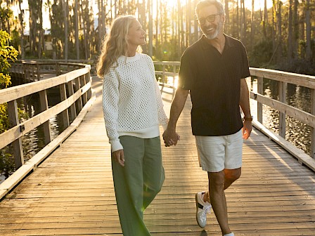 A couple is walking hand in hand on a wooden path surrounded by trees, with the sun setting behind them.