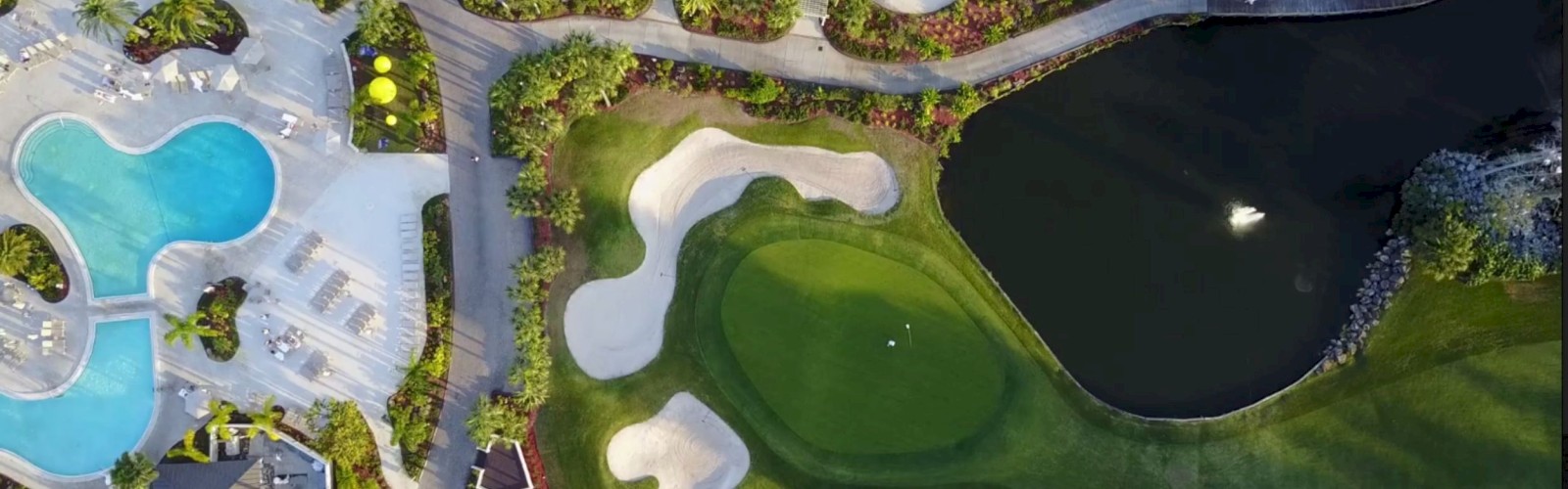 The aerial view shows a pool area, a road, a golf course, and a small lake with trees and pathways surrounding them.