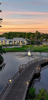 A wooden bridge crosses a lake surrounded by tall trees, leading to a house; a fountain is visible in the water under a colorful sky.