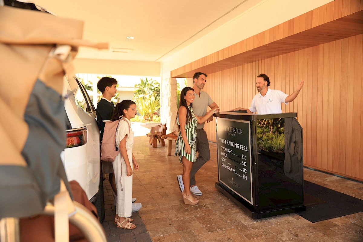 A tour group gathers around a black reception desk while a guide speaks from a podium outside a wood-paneled building.