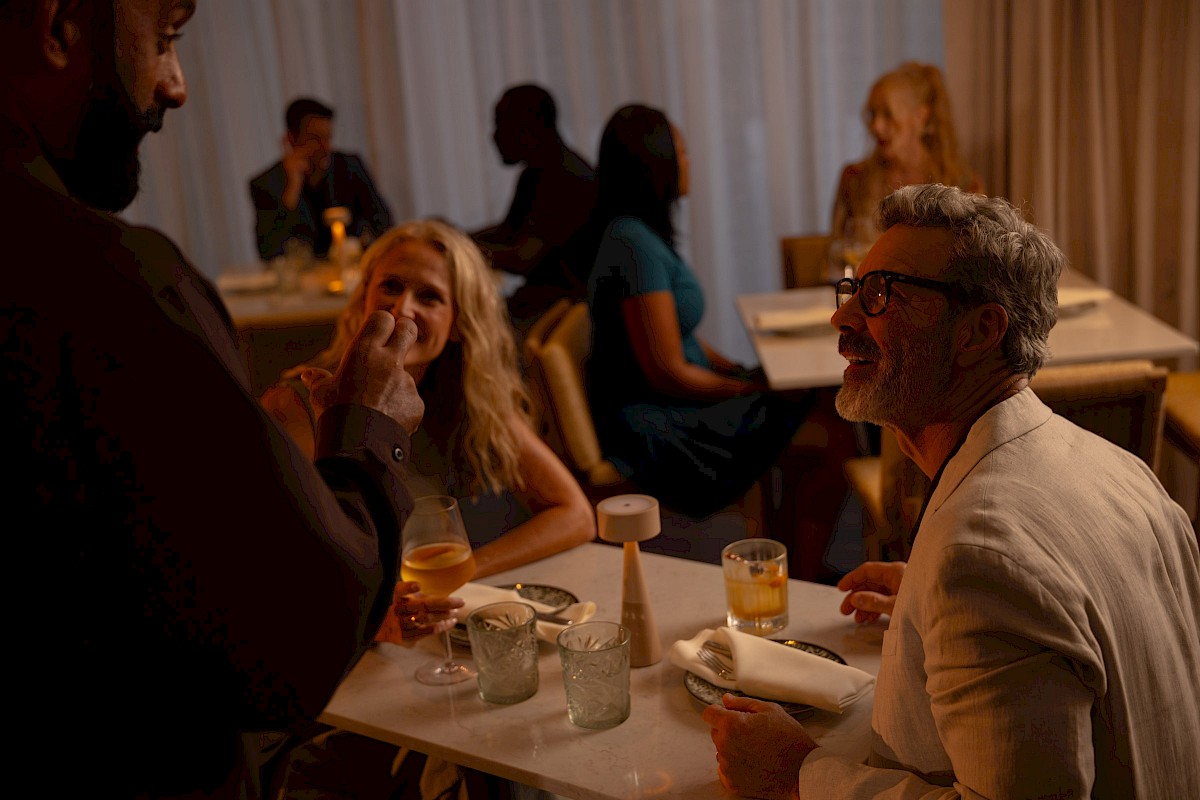 A man and a woman share a meal at a cozy restaurant, smiling and chatting with others at their table and in the background.