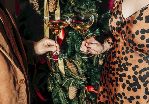 Two people clink champagne glasses in front of a decorated Christmas tree, one wearing a patterned dress.