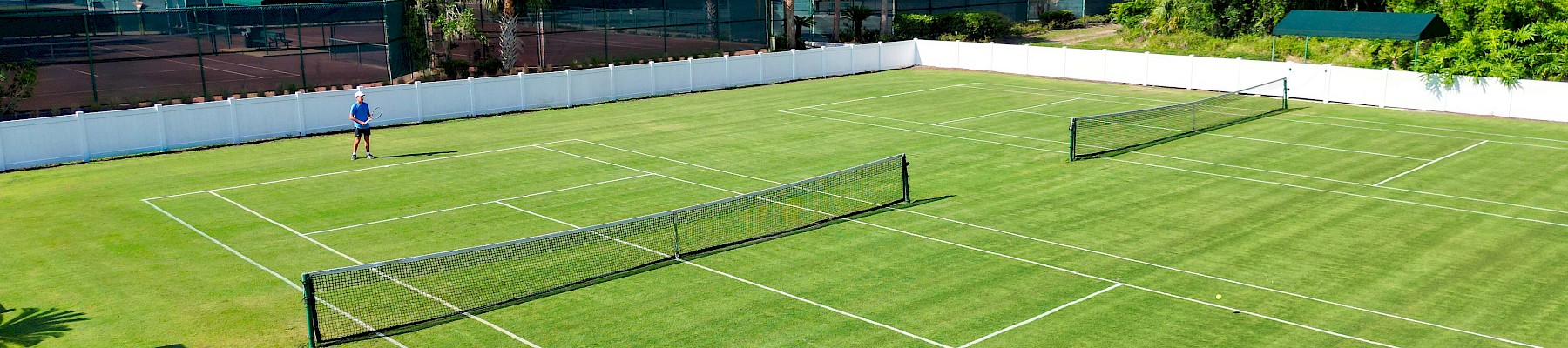 A person stands on a green grass tennis court with two nets, surrounded by trees and additional courts in the background.