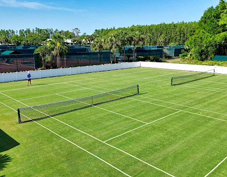 A person stands on a green grass tennis court with two nets, surrounded by trees and additional courts in the background.