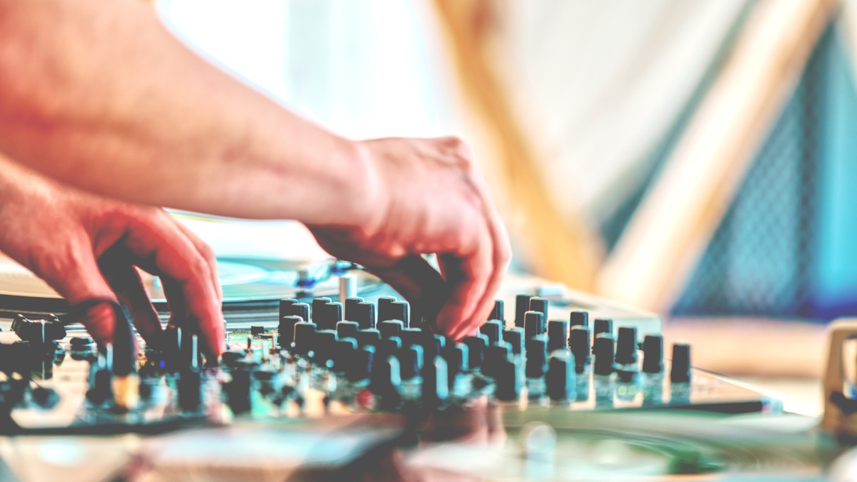 Hands are adjusting knobs on a DJ mixer, with turntables visible in a bright setting.