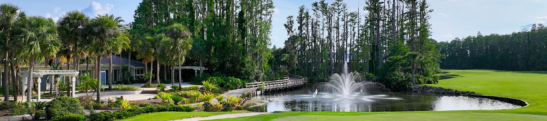 A scenic golf course with a well-kept green, water fountain, palm trees, and a clubhouse backdrop under a bright blue sky.