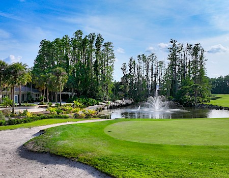 A scenic golf course with a well-kept green, water fountain, palm trees, and a clubhouse backdrop under a bright blue sky.