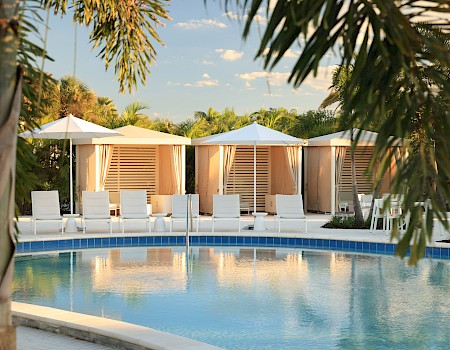 A resort pool with clear blue water, white lounge chairs, shaded cabanas, and palm trees framing a sunny, tropical scene.