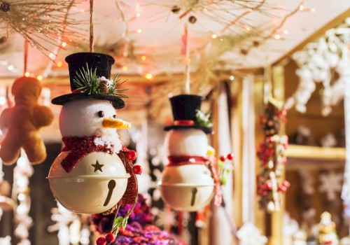 Two hanging Christmas ornaments shaped like snowmen with top hats, scarves, and bells, in a festive shop window full of holiday decorations.
