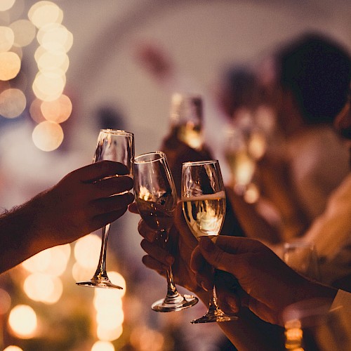 A group toasts with champagne flutes at a warmly lit celebration, clinking glasses as blurred festive lights glow in the background.