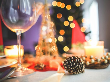 A festive table setting with wine glass, pine cone centerpiece, candles, twinkling lights, and holiday decor in a cozy dinner scene.