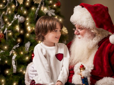 A child sits with Santa by a decorated Christmas tree, sharing a warm, festive moment.