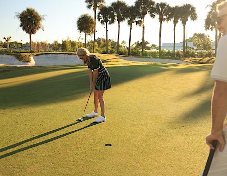 A golfer lines up a putt on a sunlit green as palm trees and fellow players watch from the edge.
