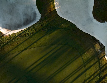 A snowy landscape with a dark green pond or puddle at the bottom, casting long shadows from nearby trees across the icy surface.