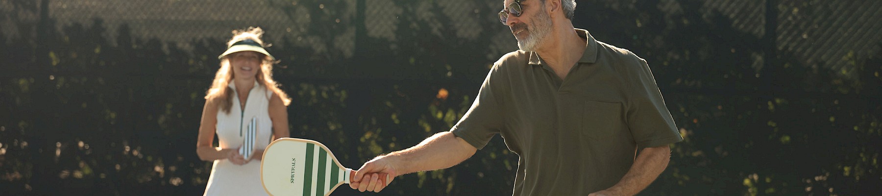 A man and a girl play tennis on an outdoor court; he’s about to serve or return, both smiling as the ball is in motion.