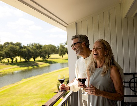 A smiling couple on a balcony overlooking a lush green landscape with a winding river, sipping red wine on a sunny day, cheers to life.