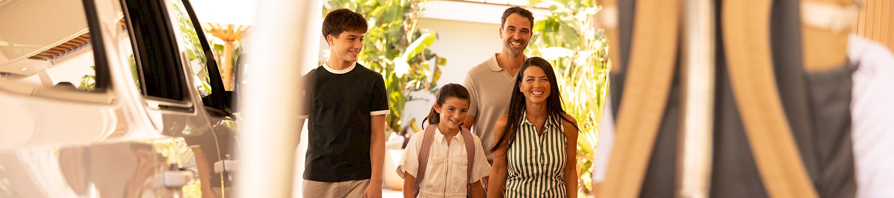 A group of people, including a child, stand near a car under a carport or hotel entrance, with a person wearing a backpack in the foreground.