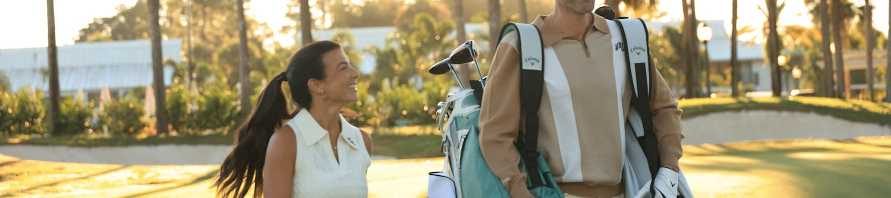 Two people, a man and a woman, walk on a sunny golf course carrying golf bags and gear, with palm trees in the background.