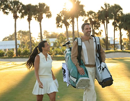 Two people, a man and a woman, walk on a sunny golf course carrying golf bags and gear, with palm trees in the background.